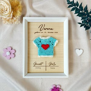 Può includere: Una cornice in legno con una maglietta da neonato blu all'uncinetto con un cuore rosso. La cornice include il nome "Verena" e la data di nascita. La targa mostra anche il peso del bambino di 3590g e l'altezza di 54cm.