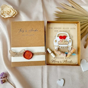 Può includere: Una scatola regalo di nozze con esterno marrone e fascia bianca, sigillata con un cuore di cera rosso. La scatola aperta rivela una carta con una grafica di auto bianca e le parole "just married". I nomi Tony & Annika e la data 26.09.2025 sono stampati.