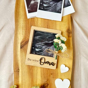 Może przedstawiać: Drewniana ramka z czarno-białym zdjęciem USG w środku. Ramka ma napis "Du wirst Oma". Na drewnianej powierzchni znajdują się dwa białe ceramiczne serca i suszone kwiaty.