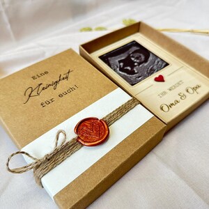 May include: A brown box with a white label that says "Eine Kleinigkeit f&uuml;r euch!" and a wax seal with a heart design. The box is open and reveals a wooden frame with a black and white ultrasound image of a baby. The frame has a red heart and the text "Ihr werdet Oma & Opa".