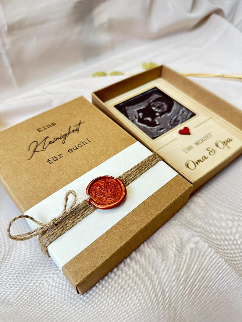 May include: A brown box with a white label that says "Eine Kleinigkeit f&uuml;r euch!" and a wax seal with a heart design. The box is open and reveals a wooden frame with a black and white ultrasound image of a baby. The frame has a red heart and the text "Ihr werdet Oma & Opa".