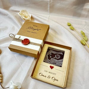 May include: A wooden box with a brown background and white text that reads "Eine Kleinigkeit f&uuml;r euch!"  The box is tied with twine and has a red wax seal.  The box is open and inside is a smaller wooden box with a photo of an ultrasound image.  The smaller box has a red heart and the text "Ihr werdet Oma & Opa".
