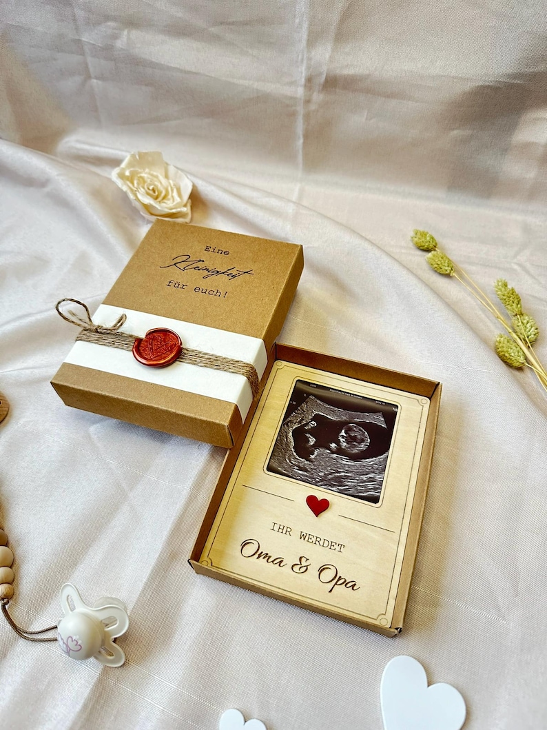 May include: A wooden box with a brown background and white text that reads "Eine Kleinigkeit f&uuml;r euch!"  The box is tied with twine and has a red wax seal.  The box is open and inside is a smaller wooden box with a photo of an ultrasound image.  The smaller box has a red heart and the text "Ihr werdet Oma & Opa".