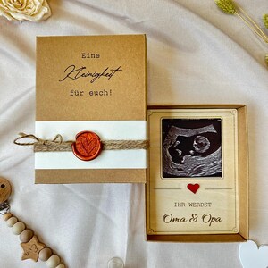 May include: A brown box with a white label that says "Eine Kleinigkeit f&uuml;r euch!" and a red wax seal. A wooden frame with a black and white ultrasound image and the text "Ihr werdet Oma & Opa" in red.