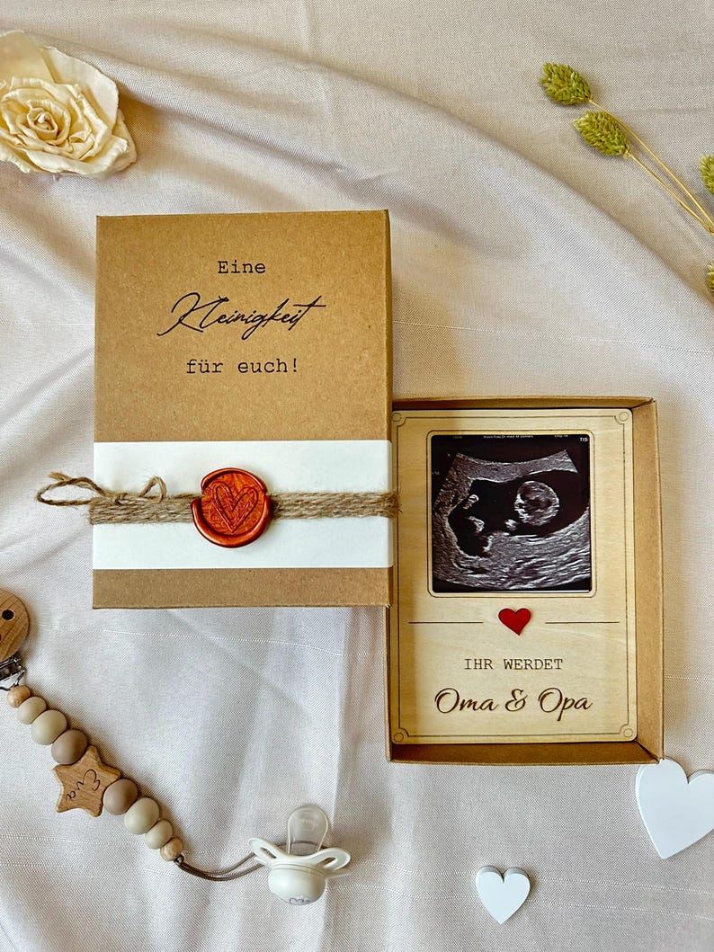 May include: A brown box with a white label that says "Eine Kleinigkeit f&uuml;r euch!" and a red wax seal. A wooden frame with a black and white ultrasound image and the text "Ihr werdet Oma & Opa" in red.