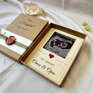 May include: A wooden box with a brown paper exterior and a red heart. The box contains a card with a black and white ultrasound image and the text "Ihr werdet Oma & Opa".