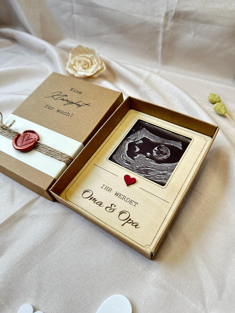 May include: A wooden box with a brown paper exterior and a red heart. The box contains a card with a black and white ultrasound image and the text "Ihr werdet Oma & Opa".