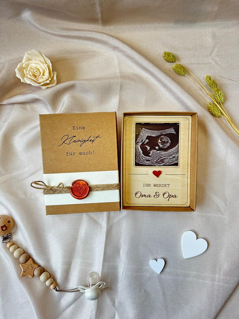 May include: A brown box with a white label that says "Eine Kleinigkeit fur euch!" and a red wax seal. Next to it is a wooden box with a brown frame and a black and white ultrasound image inside. The box has a red heart and the text "Ihr werdet Oma & Opa".