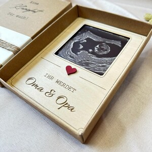 May include: A brown box with a white interior. The box is open and contains a small ultrasound image of a baby. The image is framed with a white border and a red heart. The text "Ihr werdet Oma & Opa" is written on the box.
