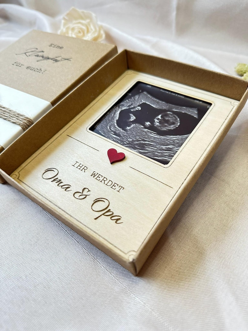May include: A brown box with a white interior. The box is open and contains a small ultrasound image of a baby. The image is framed with a white border and a red heart. The text "Ihr werdet Oma & Opa" is written on the box.