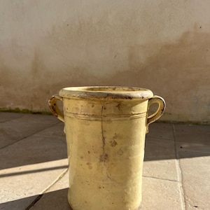 Puede incluir: Una maceta cilíndrica de cerámica amarilla antigua con dos asas pequeñas. La maceta tiene un aspecto ligeramente desgastado con grietas e imperfecciones visibles, lo que sugiere edad y uso. La maceta es probablemente para fines decorativos o funcionales.