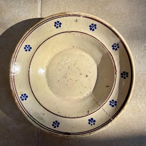 Peut inclure: Une assiette ancienne de couleur crème, ornée d'un bord rouge foncé et de motifs floraux bleus. L'assiette, de forme ronde, présente une zone centrale surélevée et une fissure. Le diamètre est d'environ 25 cm.