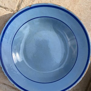 Op de afbeelding: Een lichtblauwe keramische plaat met een donkerblauwe rand en een donkerblauwe concentrische cirkel. De plaat heeft een licht versleten uiterlijk met zichtbare scheuren en onvolkomenheden. De plaat is rond.