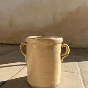 Puede incluir: Una olla de cerámica de color crema, de forma cilíndrica, con dos pequeñas asas. La olla tiene un borde ligeramente ensanchado y una superficie texturizada. Probablemente se utiliza para almacenamiento o como objeto decorativo.