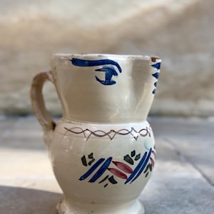 Peut inclure: Pichet en céramique crème avec une anse, décoré de motifs abstraits bleus, rouges et verts. Le pichet présente un œil bleu peint sur le devant et un bord décoratif.