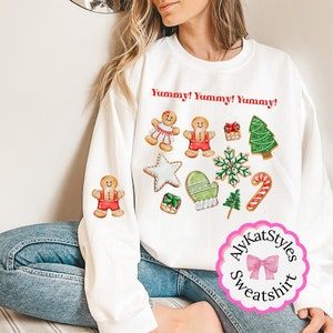 May include: White sweatshirt with a Christmas gingerbread cookie theme. The text "Yummy! Yummy! Yummy!" is printed on the front of the sweatshirt. The sweatshirt has a pink and white label with the text "AlyKatStyles Sweatshirt".