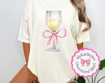 May include: A white oversized t-shirt with a graphic of a glass of white wine tied with a pink ribbon. The text "AlyKatStyles Oversized Shirt" is in a pink circle with a pink bow.