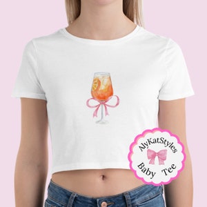 Può includere: Un top corto bianco con un nastro rosa attorno a un bicchiere di succo d'arancia con una fetta d'arancia al suo interno. Il testo "AlyKatStyles Baby Tee" è su un cerchio rosa.