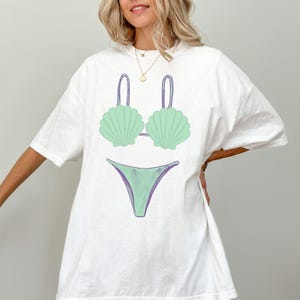 May include: White t-shirt featuring a seafoam green bikini design. The bikini top has a seashell shape, and the bottom is a simple V-shape. The design has purple accents. The shirt is a casual, oversized fit.
