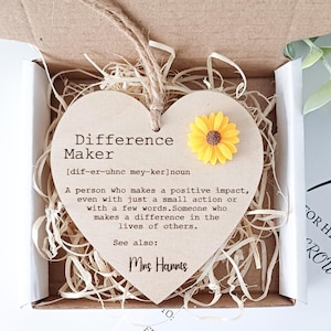 May include: A wooden heart-shaped plaque with the word "Difference Maker" defined as "a person who makes a positive impact, even with just a small action or with a few words." The plaque has a yellow sunflower with a brown center. The plaque is in a small box with wood shavings.