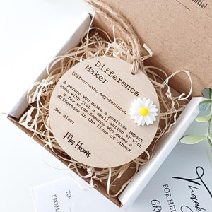 May include: A round wooden tag with the words "Difference Maker" and a definition of the word. The tag is decorated with a white daisy with a yellow centre. The tag is in a small box with wood shavings.