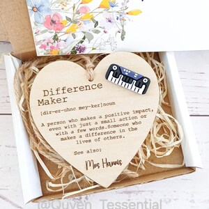 Puede incluir: Una caja de regalo con una placa de madera en forma de corazón que dice "Difference Maker" y un pequeño pin de teclado. La caja también contiene una tarjeta con diseños florales y las palabras "Thank you".