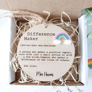 Puede incluir: Un letrero redondo de madera con las palabras "Difference Maker" y una definición de la palabra. El letrero también incluye un arcoíris con nubes y el nombre "Mrs. Hannis".