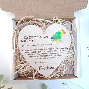 Puede incluir: Adorno de madera en forma de corazón con una definición de "Difference Maker" y un pequeño loro pintado. El adorno está en una caja con papel triturado y una tarjeta de agradecimiento que dice "¡Gracias por ayudarme a crecer!"