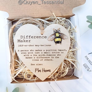 May include: Wooden heart-shaped plaque with a bee illustration and the text "Difference Maker" with a definition and the name "Mrs. Hannis". The plaque is in a small box with brown shredded paper.