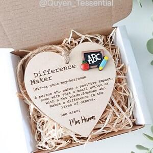 Puede incluir: Adorno de madera en forma de corazón con las palabras "Difference Maker" y una definición. El adorno incluye una pequeña pizarra, una manzana y un lápiz. Se presenta en una caja de regalo con paja.