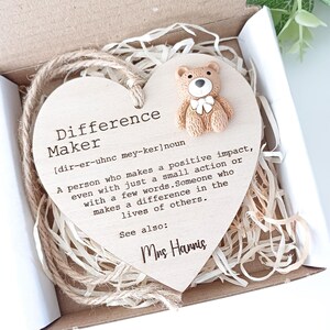 Puede incluir: Una placa de madera en forma de corazón con la palabra "Difference Maker" definida como "una persona que tiene un impacto positivo, incluso con una pequeña acción o con unas pocas palabras". La placa también incluye el texto "Ver también: Sra. Harris" y una pequeña figurita de oso de peluche marrón.