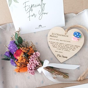 Puede incluir: Un juego de regalo con un pequeño ramo de flores secas en tonos morados, naranjas y rosas, atado con una cinta blanca. Incluye una placa de madera en forma de corazón con el texto "Difference Maker" y una tarjeta que dice "Especially For You".