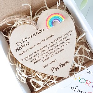May include: A heart-shaped wooden plaque with the words "Difference Maker" and a definition printed on it. A small, colorful rainbow decoration is attached. The plaque is presented in a box with straw.