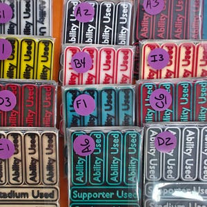 May include: A collection of rectangular game tokens in clear plastic packaging. Each token has the words "Ability Used" printed on it. The tokens are in various colors, including yellow, red, and teal, with purple stickers.