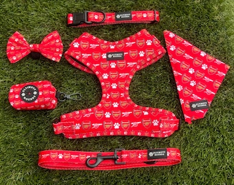 May include: A red dog harness with a white paw print and Arsenal logo pattern. The harness is part of a set that includes a matching leash, bow tie, poop bag holder, and bandana. The set is branded with "Ultimutt Bows".