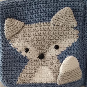 May include: A handmade crocheted item with a fox motif. The fox is depicted in shades of grey and white, with black button eyes, on a blue background. The item has a textured, handcrafted appearance.