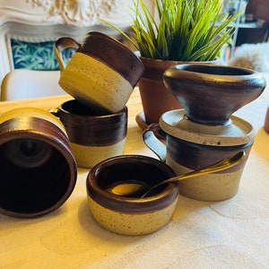 May include: Set of four brown and beige ceramic mugs with a speckled glaze. The mugs have a rounded shape and a handle. There is also a small brown and beige ceramic bowl with a speckled glaze and a gold spoon inside.
