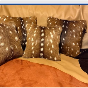 May include: A set of four square throw pillows covered in brown animal hide with a distinct dark brown central stripe and scattered white spots. The pillows feature a textured, fur-like surface and are arranged in a group on a smooth tan surface.