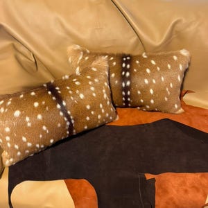 Puede incluir: Dos cojines rectangulares decorativos con un patrón moteado marrón y blanco, que recuerda a la piel de ciervo. Se colocan sobre una superficie de cuero marrón y beige, creando una estética rústica. Estos cojines son un elemento de decoración del hogar.