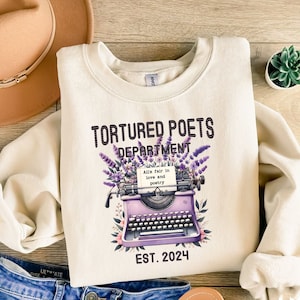 May include: A cream colored sweatshirt with a purple typewriter and lavender flowers. The text "Tortured Poets Department" is above the typewriter and the text "All's fair in love and poetry" is on a piece of paper in the typewriter. The text "EST. 2024" is below the typewriter.