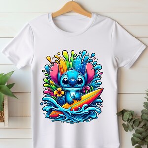 May include: A white t-shirt with a colorful graphic of a blue cartoon character surfing on a yellow surfboard. The character has large eyes and a big smile. The background is filled with colorful splashes of water and flowers.