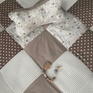 Könnte beinhalten: Eine braune und weiße Patchwork-Babydecke mit braunem Punkte-Muster, weißem strukturiertem Muster und einem Muster aus Lamas. Ein Holzspielzeug mit braunem und weißem Muster liegt auf der Decke.