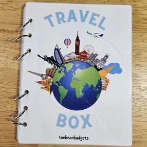 May include: A white travel journal with a blue and white illustration of a globe surrounded by famous landmarks. The text "TRAVEL BOX" is in blue letters above the globe and "toebeanbudgets" is in black letters below the globe.