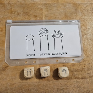 May include: A clear plastic pouch containing a white card with black line drawings of rock, paper, and scissors represented by paw shapes. Below the card are three wooden dice, each with a different paw illustration.