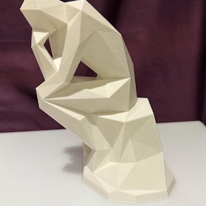 Puede incluir: Una escultura geométrica de color crema de una persona en una pose sentada y contemplativa. La forma de la figura está construida a partir de planos triangulares y cuadriláteros interconectados, creando una estética moderna y abstracta. La escultura mide unos 20 cm de altura.