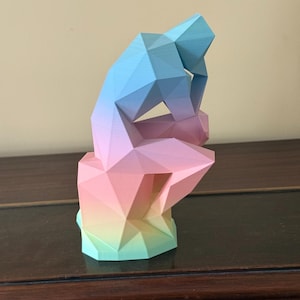 Puede incluir: Una escultura geométrica de una persona en pose pensativa, representada en un degradado de colores. La escultura transiciona del azul al rosa, luego al amarillo y al verde. La figura está sentada sobre una base.