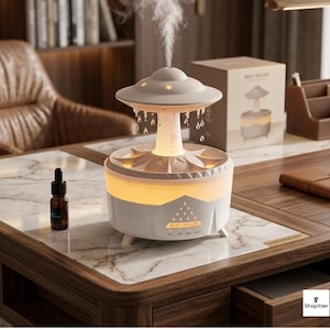 Peut inclure: Un diffuseur d'huiles essentielles blanc et gris au design unique, avec un dessus en forme d'OVNI avec des découpes en forme d'étoiles. La base présente un motif géométrique et l'inscription "BEST-SELLER". Une petite bouteille d'huile essentielle est également visible.