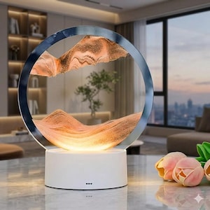 Può includere: Lampada decorativa con sabbia artistica e cornice circolare in vetro. La cornice contiene strati di sabbia arancione e marrone, creando un paesaggio montuoso. La lampada poggia su una base bianca con luce calda.