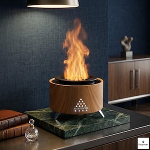 May include: A wooden-effect tabletop fireplace with a black top and silver legs, emitting a bright orange flame. The fireplace sits on a green marble base. A small glass perfume bottle and books are in the foreground.