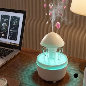 Puede incluir: Un humidificador blanco con forma de seta, base de luz turquesa y detalles transparentes similares a tentáculos. Emite un chorro vertical de vapor, mientras que unas gotas rosas parecen flotar en el aire sobre la parte superior.